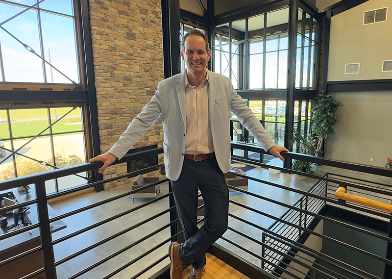 A man in a light blue blazer stands on the landing of a staircase with an open lobby behind him